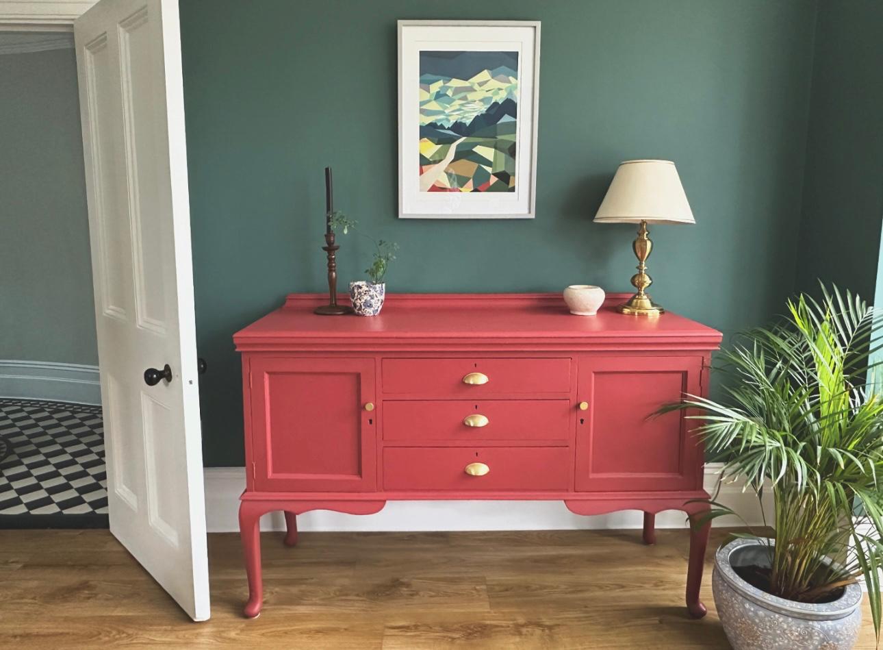 Vibrant coral sideboard against a deep green wall with abstract art, showcasing Emsworth interior design.